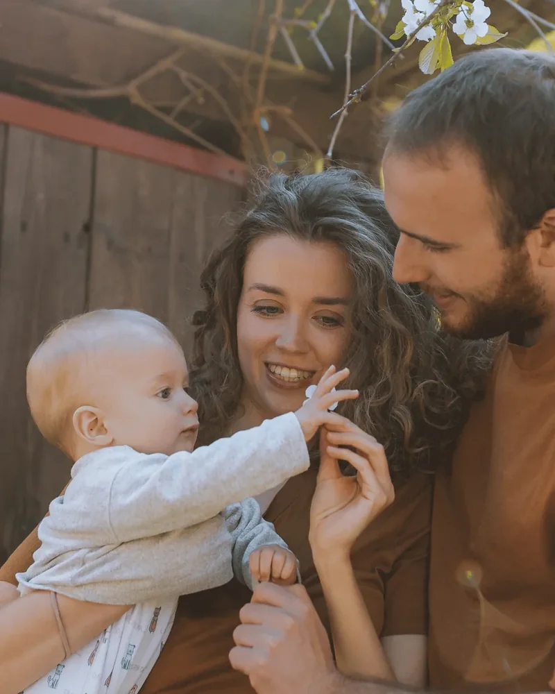 A mother and father outside a barn in spring, their baby reaching up to touch her hand